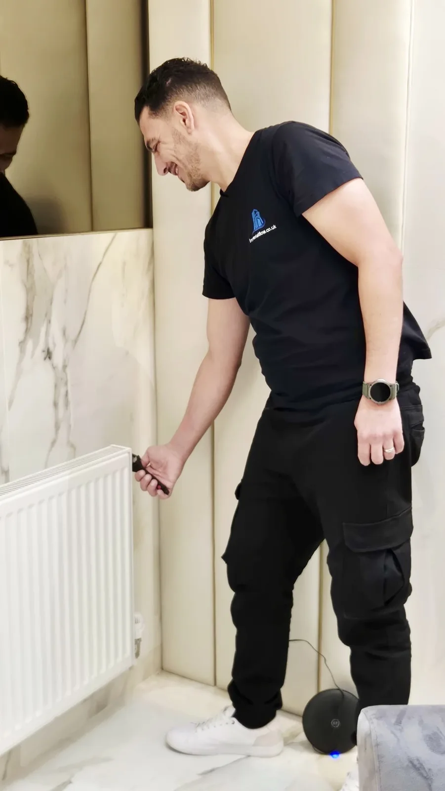 Imperial Flow adjusting a radiator valve on a marble-tiled wall, under brass mirrored cabinets.