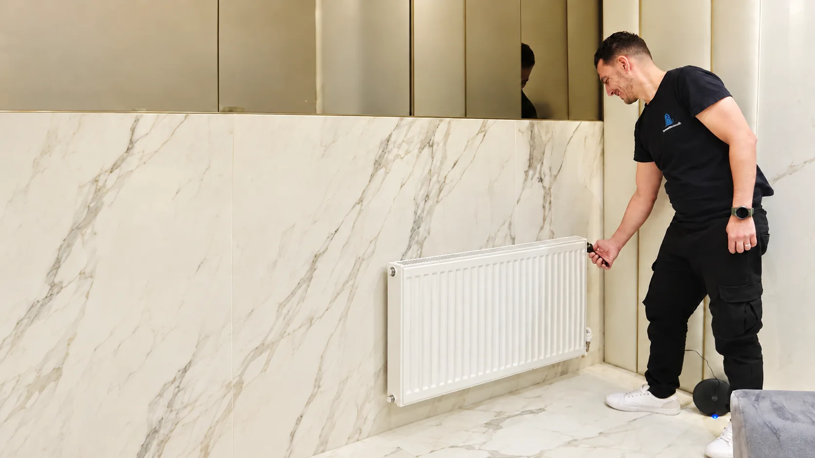 Imperial Flow adjusting a radiator valve on a marble-tiled wall, under brass mirrored cabinets.